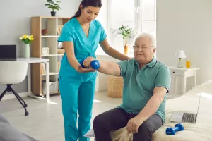Älterer Mann führt Physiotherapie-Übungen mit einer Hantel durch. Krankenschwester-Physiotherapeutin hilft ihm dabei.